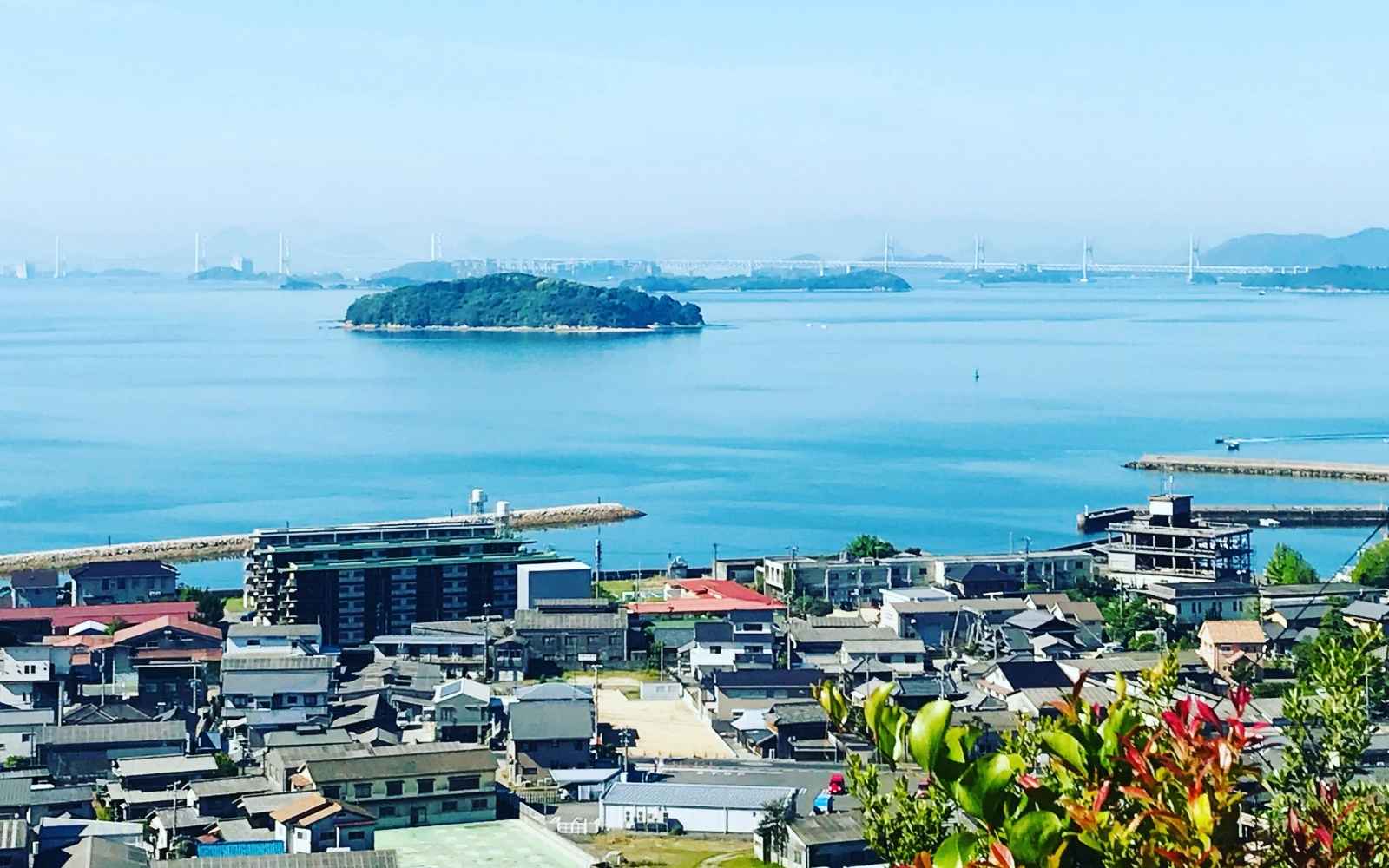 A view of Karakoto Kojima, Kurashiki the town where we make our fabric. Woven in Japan at our family business. A  small town with many buildings nestled in a small area.  In the background the blue Setouchi Sea and the Seto Ohashi Bridge can be seen.  